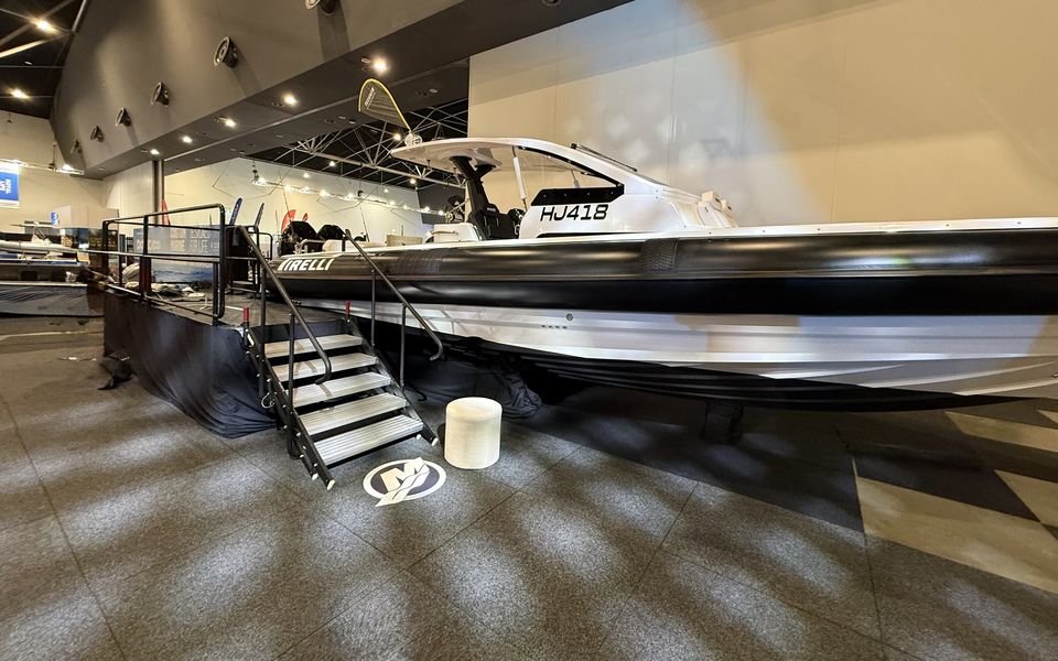 Boat displayed on a raised stage with black skirting and access stairs at an indoor boat show