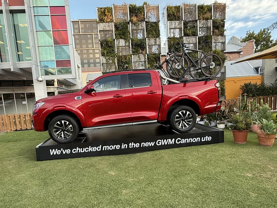 Red ute with bikes on the tray on a raised display platform on a lawn, with a green wall behind