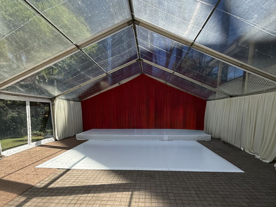 White stage and white dance floor inside a clear-roof marquee with a red curtain backdrop