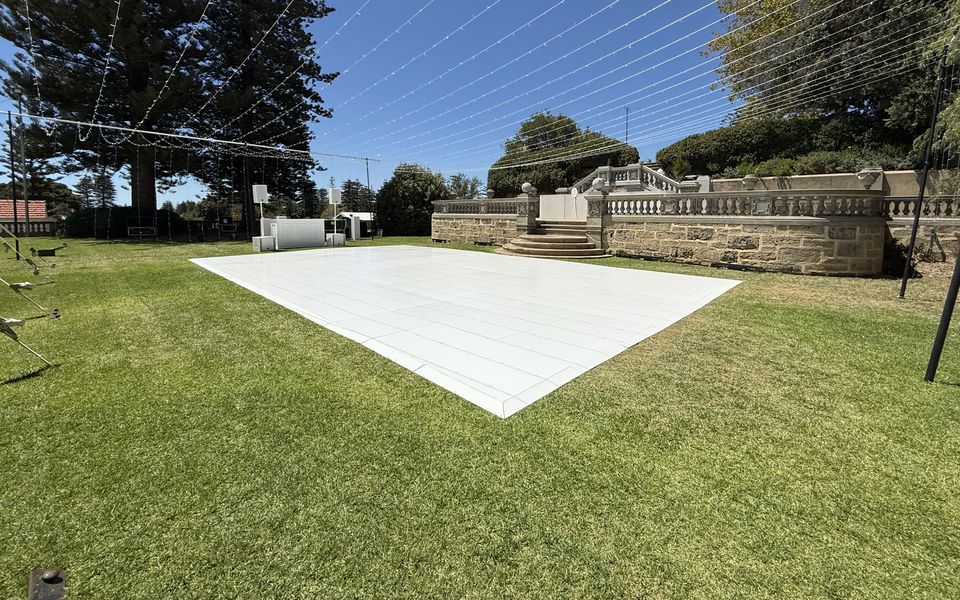 White dance floor laid on a garden lawn under festoon wires, with a stone balustrade and pine trees behind
