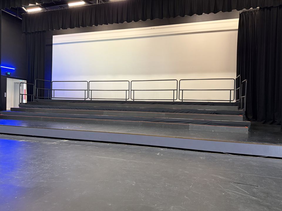 Black tiered stage with handrails, set up for a graduation or choir in front of a white backdrop
